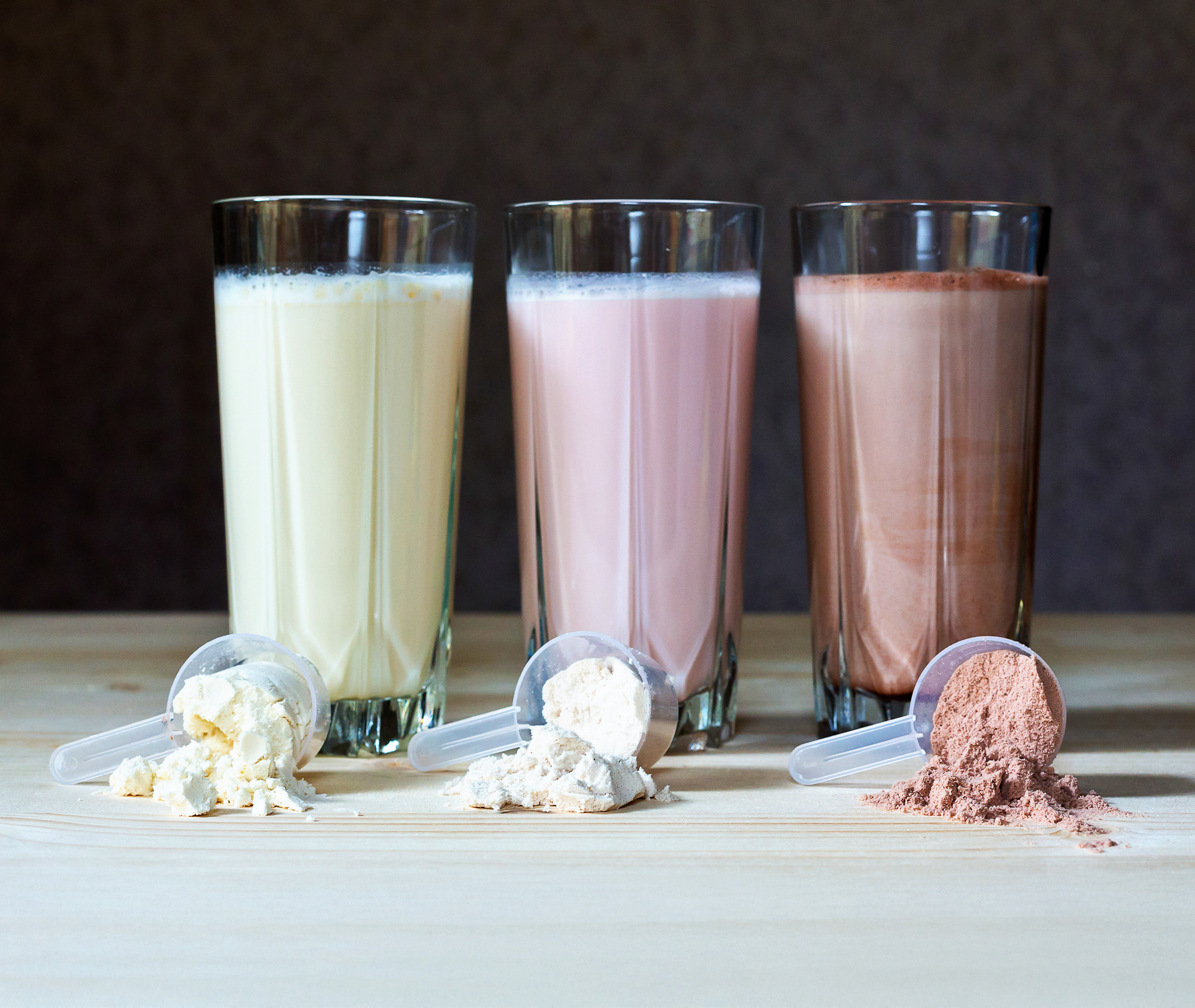 Drei Gläser mit Proteinshakes und jeweils ein Scoop mit Pulver im Vordergrund